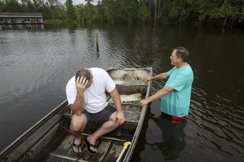 A man sits in a boat floating on flood waters beside another man.
