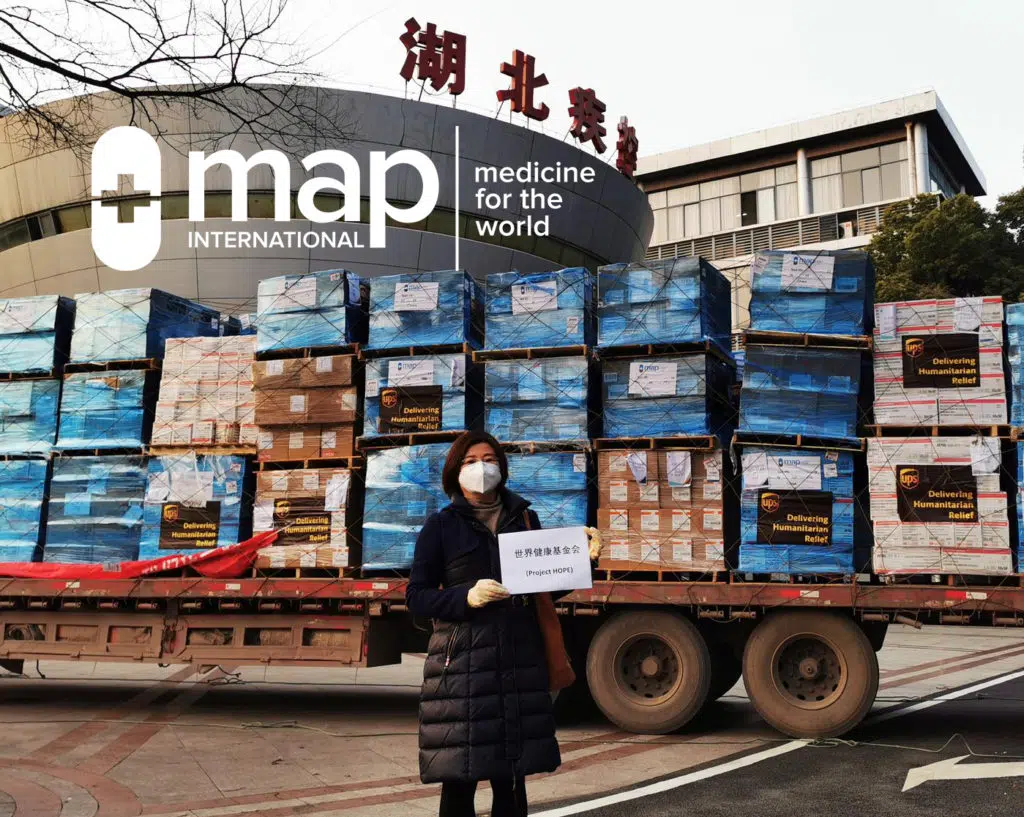 A woman stands holding a Project HOPE sign in front of MAP International supplies shipped by UPS in China.