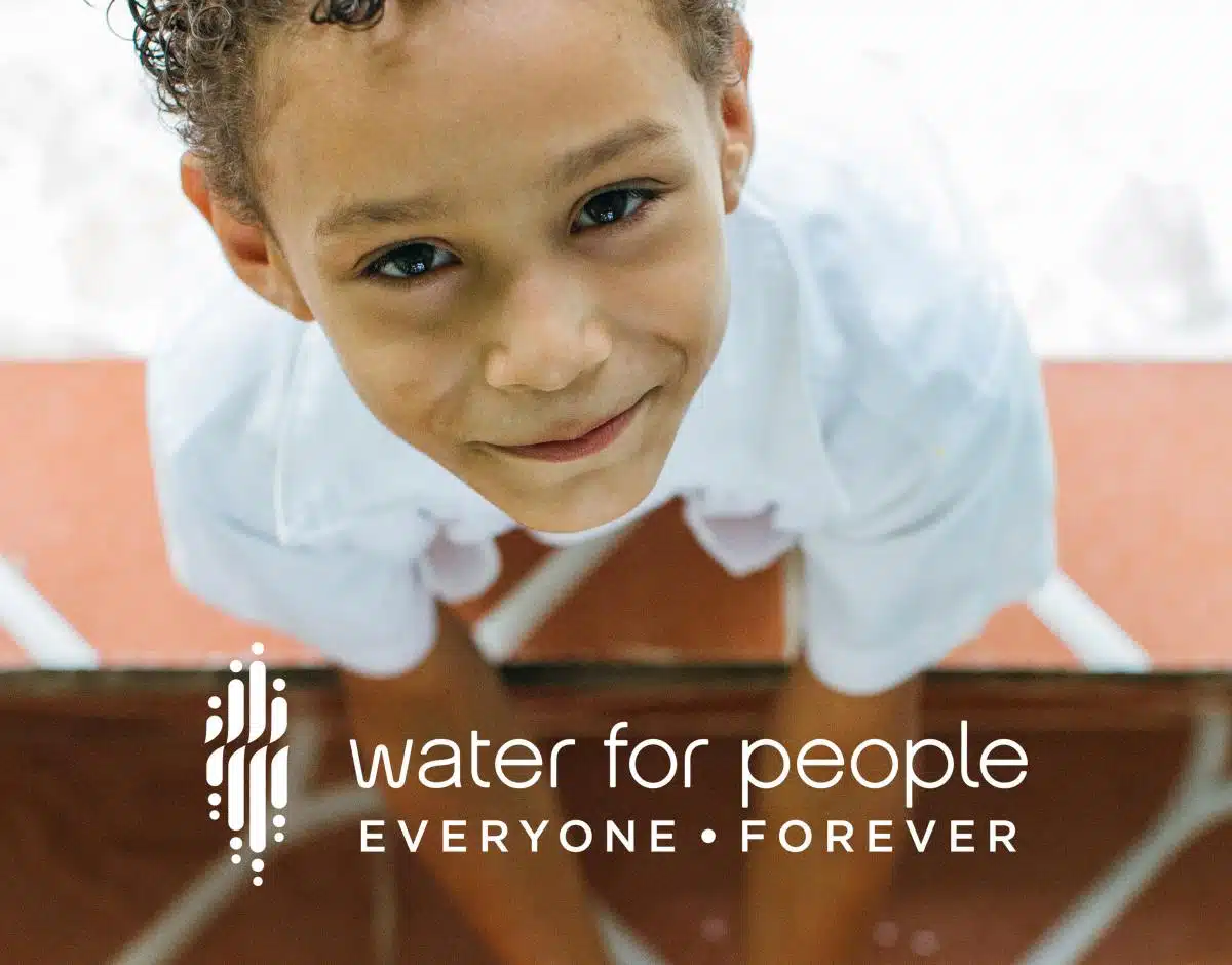 Water For People logo over photo of a boy looking up while he washes his hands