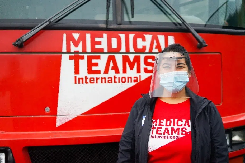 A woman stands in front of a red Medical Teams International vehicle wearing a Medical Teams International shirt. .