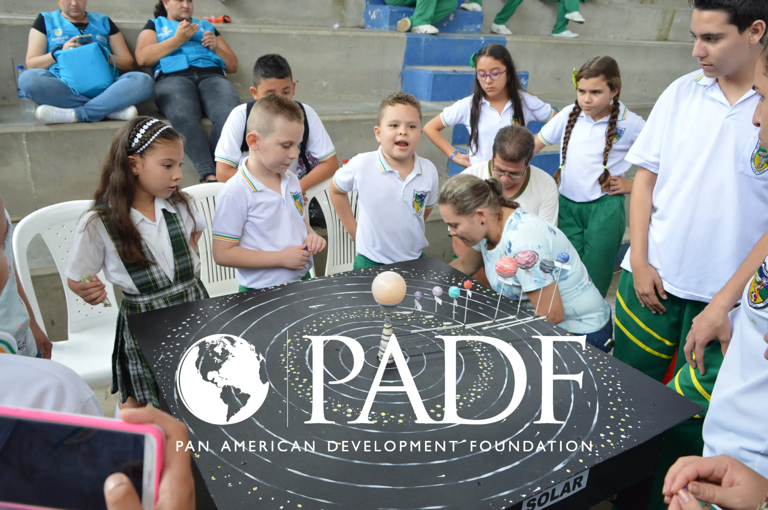 A group of school children in Colombia gather around a planetary model. Pan American Development Foundation logo.