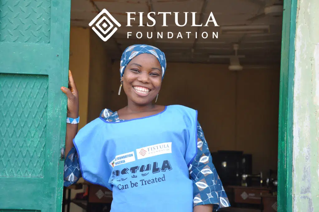 A woman wearing a Fistula Foundation shirt stands in a doorway and smiles.