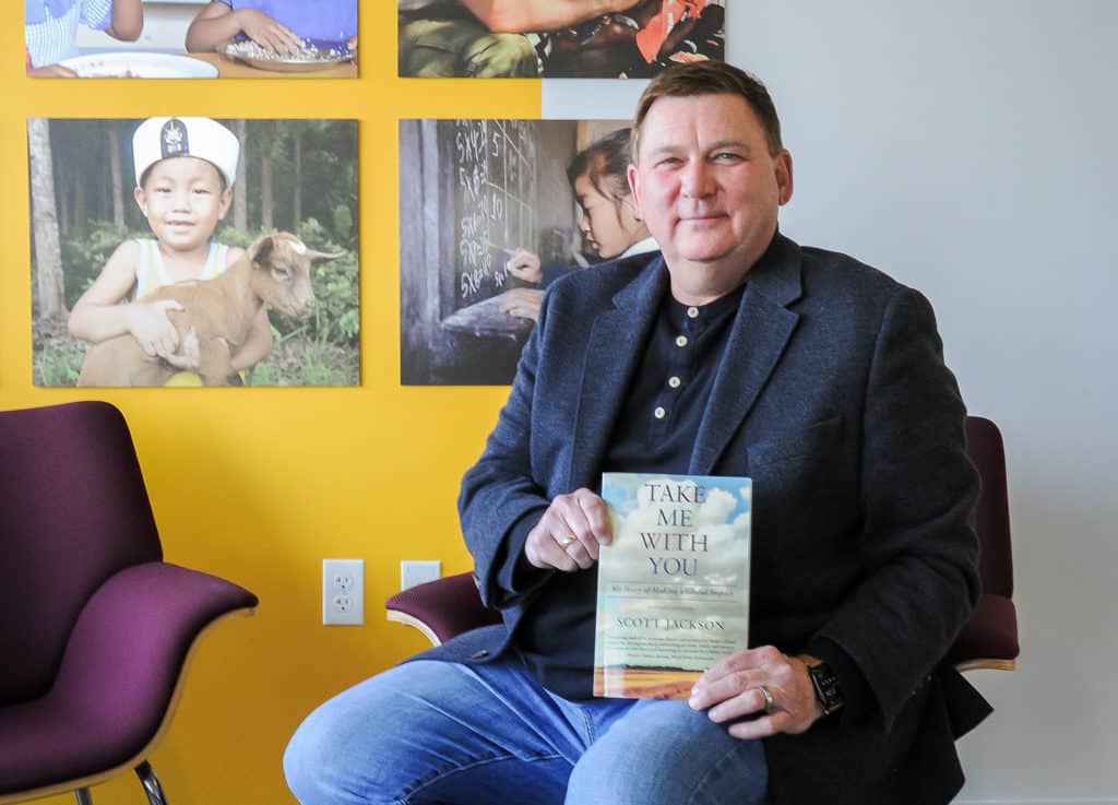 Scott Jackson sitting in a chair at Global Impact headquarters in front of charity photos holding a copy of his new book.