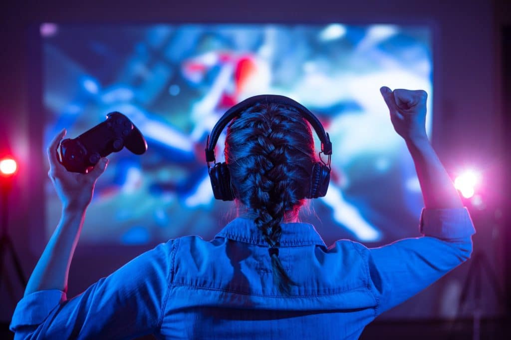 A girl facing away from the camera holding a Playstation controller sitting in front of a screen displaying a game and cheering with her arms in the air.