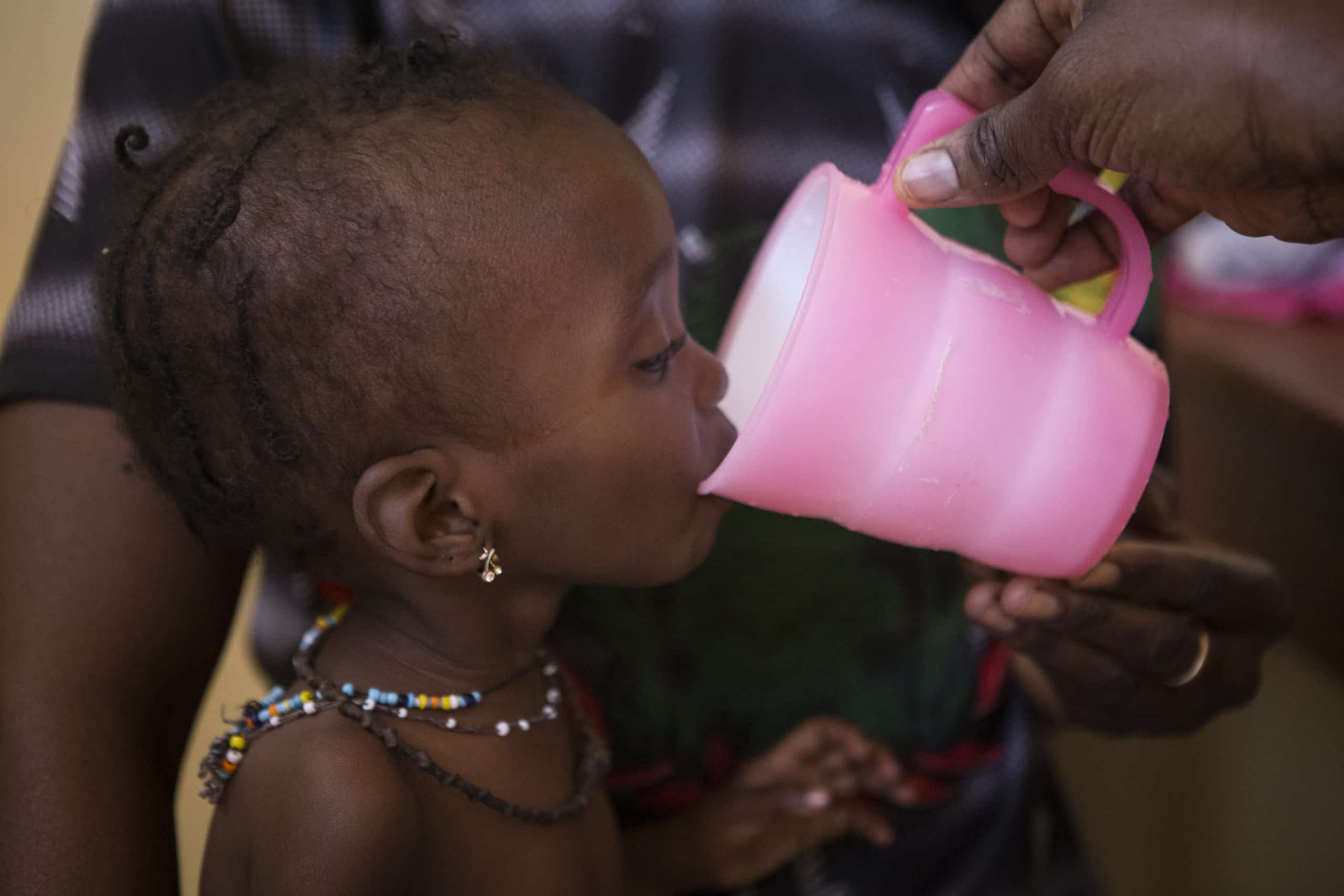 A black child drinking water from the pink cup that a black person is holding it