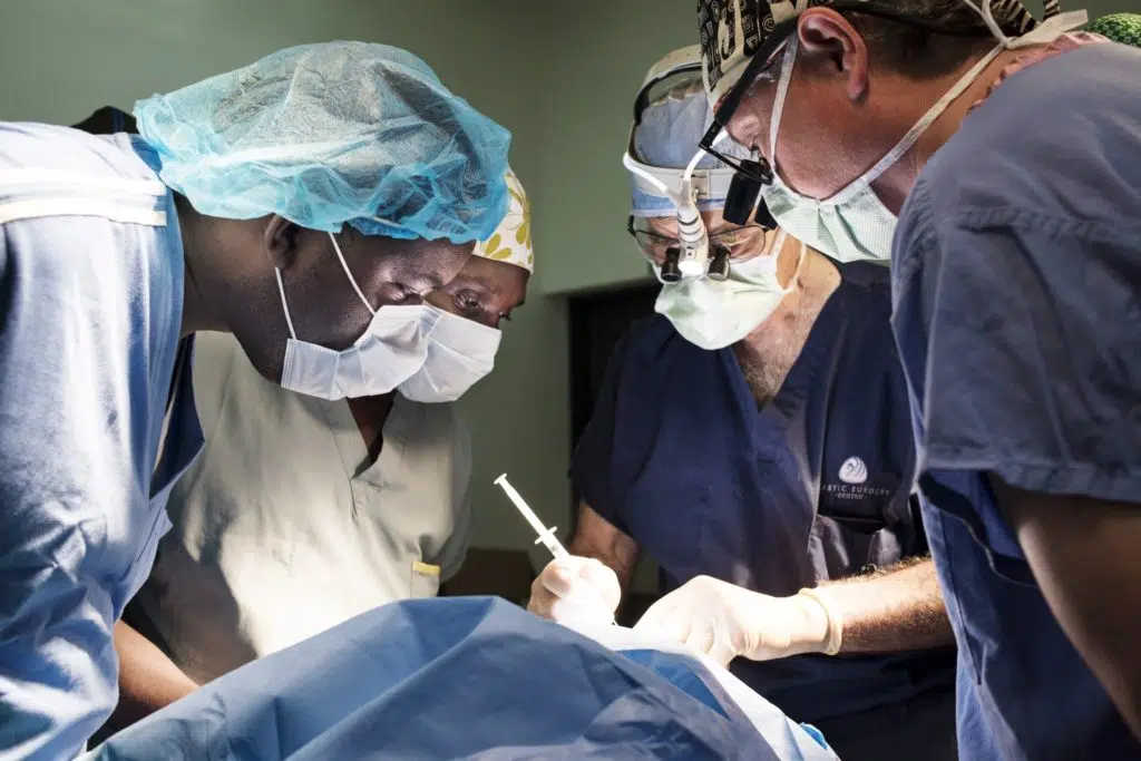 A surgeon performing a surgery while a white male technican and two black (one male and one female) technicans are observing