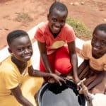 Three kids smiling up around a bucket of water