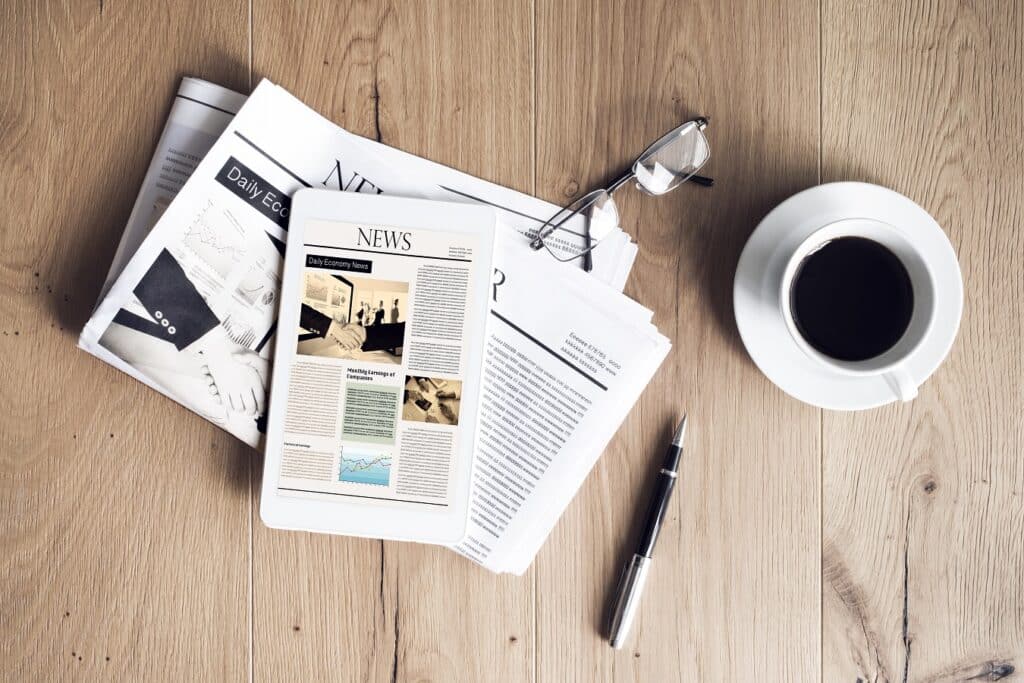 A desk with news papers, a coffee mug, glasses and a pen.