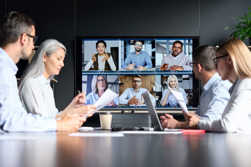Online video conference in a meeting room with people sitting in the office and colleagues on the big screen monitor.