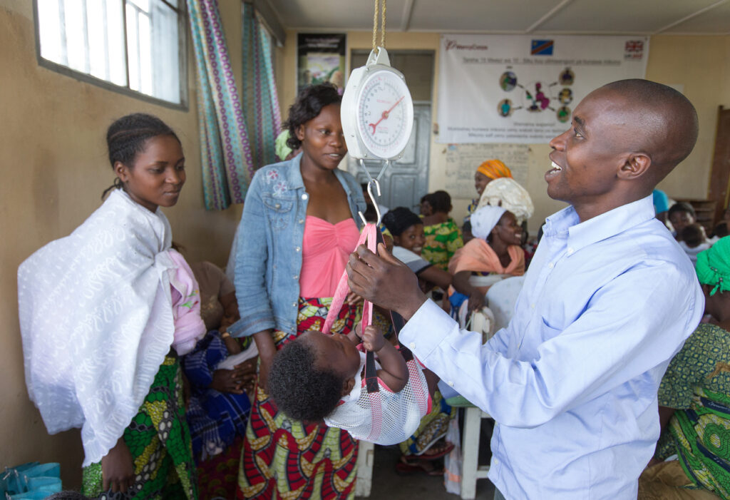 Lukogho Mukandirwa Vianney weighs Flaura Numbere while her mother Lydia Kahambu (center) looks on at the Majengo Health Center in Goma, Democratic Republic of Congo. Photo by Mike DuBose, UMNS
