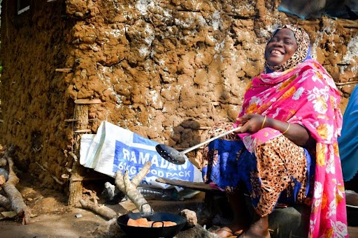 Aichata smiling and cooking over a fire, with a Ramadan Food Pack in the background.